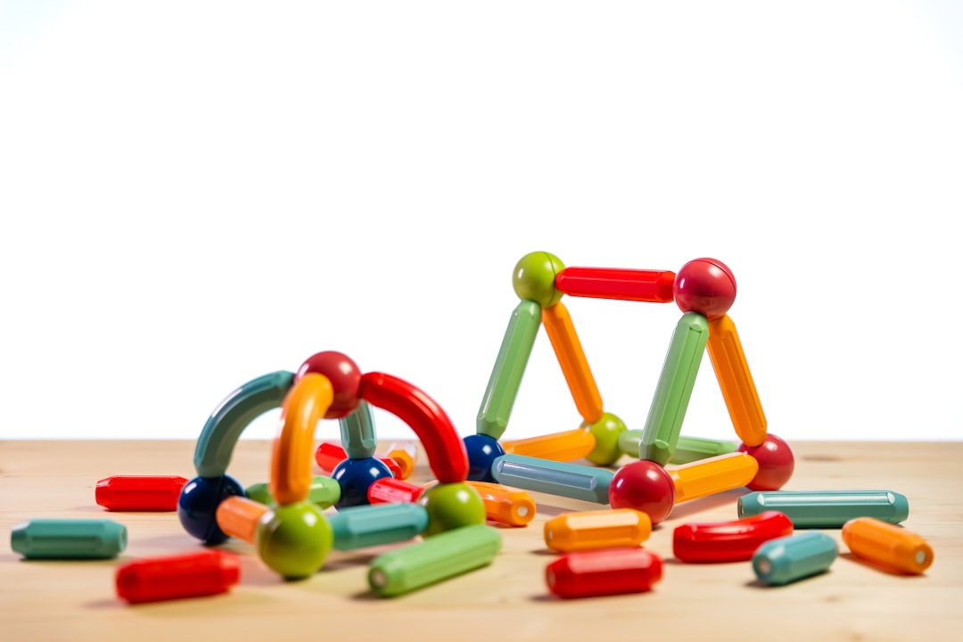 A close-up shot of colorful magnetic building blocks assembled on a light wooden surface against a white background. The blocks, which consist of spheres and rods, are connected to form small abstract structures, including an arch and a triangle shape.