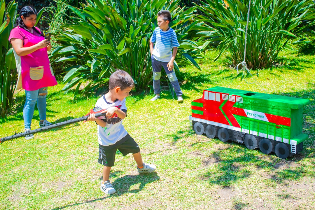 Young boy hitting a colorful train-shaped piñata outdoors while another child and a woman watch in a sunny garden setting
