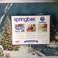 Springbok The Falling Snow 500 Piece Jigsaw Puzzle For Adults Features A Holiday Cabin Glowing In The Snow With A Decorated Christmas Tree Beside It