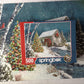 Springbok The Falling Snow 500 Piece Jigsaw Puzzle For Adults Features A Holiday Cabin Glowing In The Snow With A Decorated Christmas Tree Beside It